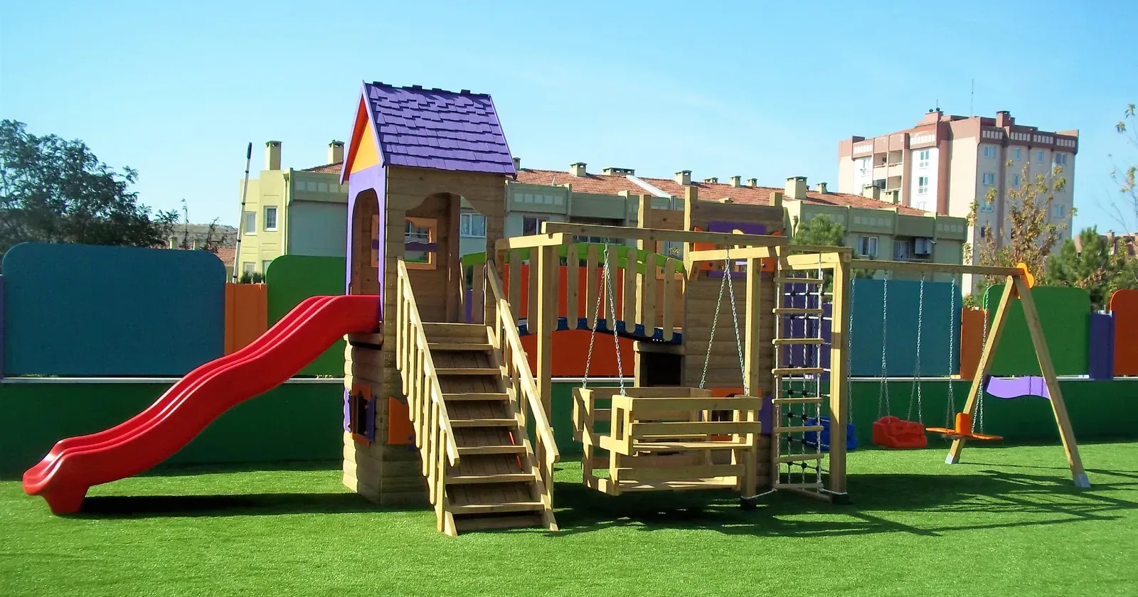 Anaokulu bahçesinde kurulmuş ahşap oyun parkı; kaydırak, salıncak ve tırmanma bölümleri