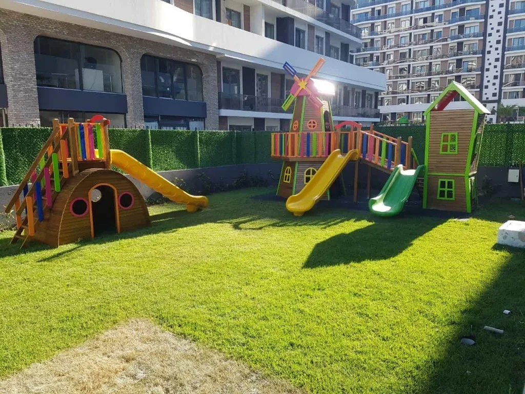 Anaokulu bahçesinde renkli ahşap oyun parkı — kaydıraklar, yel değirmeni ve oyun kulübesi